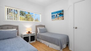 A bedroom with two beds, a nightstand with a lamp, a painting of a beach scene, and a window with blinds.