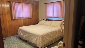 Ein Schlafzimmer mit einem großen Bett, zwei Vorhängen, einem Holzkopfteil, einem Nachttisch mit Lampe und einem Sessel.