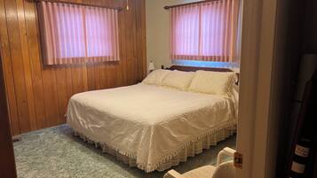 A bedroom with a large bed, two windows with curtains, a wooden headboard, a bedside table with a lamp, and a chair.