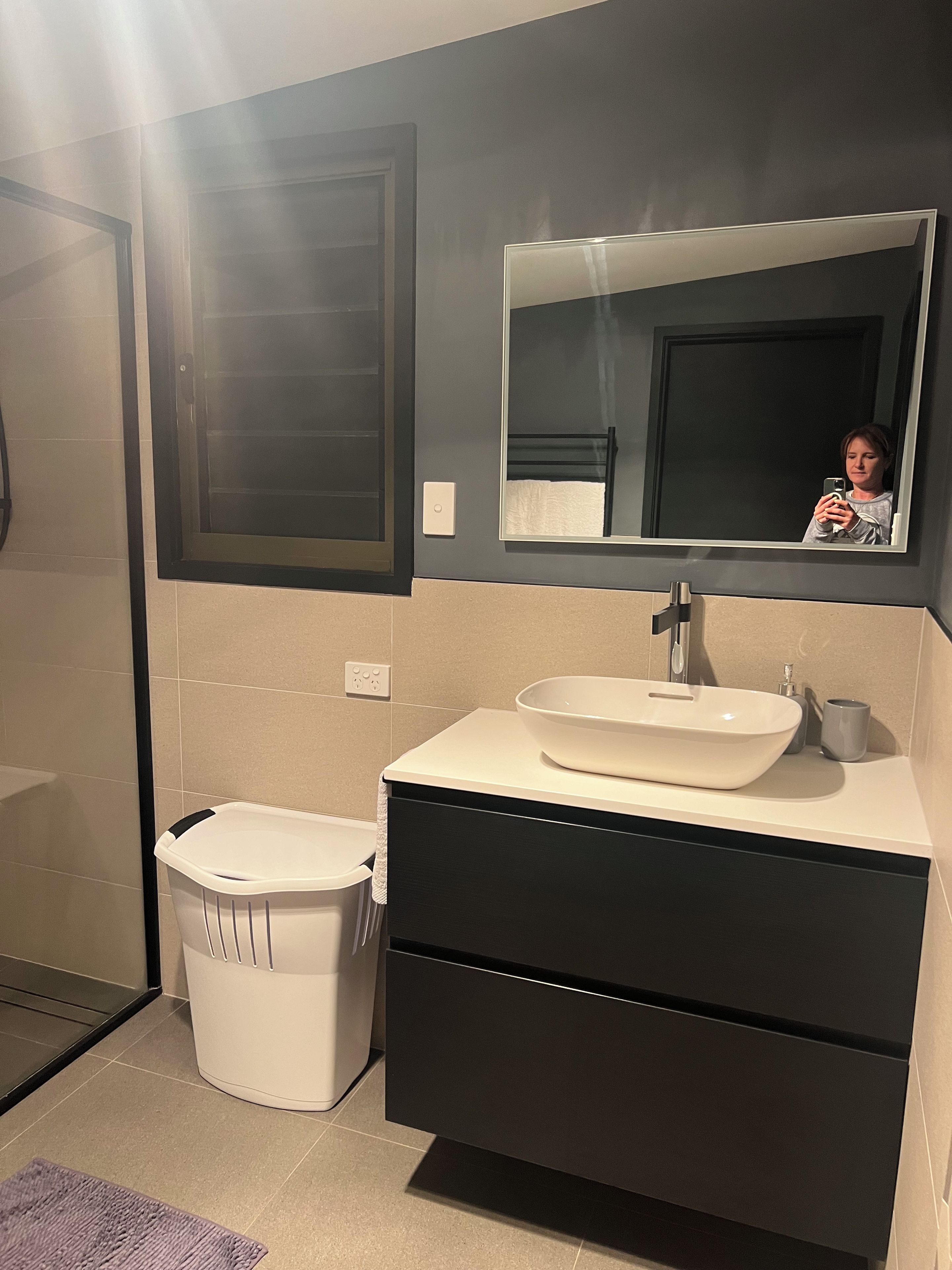 Une salle de bain avec un lavabo blanc, des meubles de rangement de couleur foncée et un miroir au-dessus.