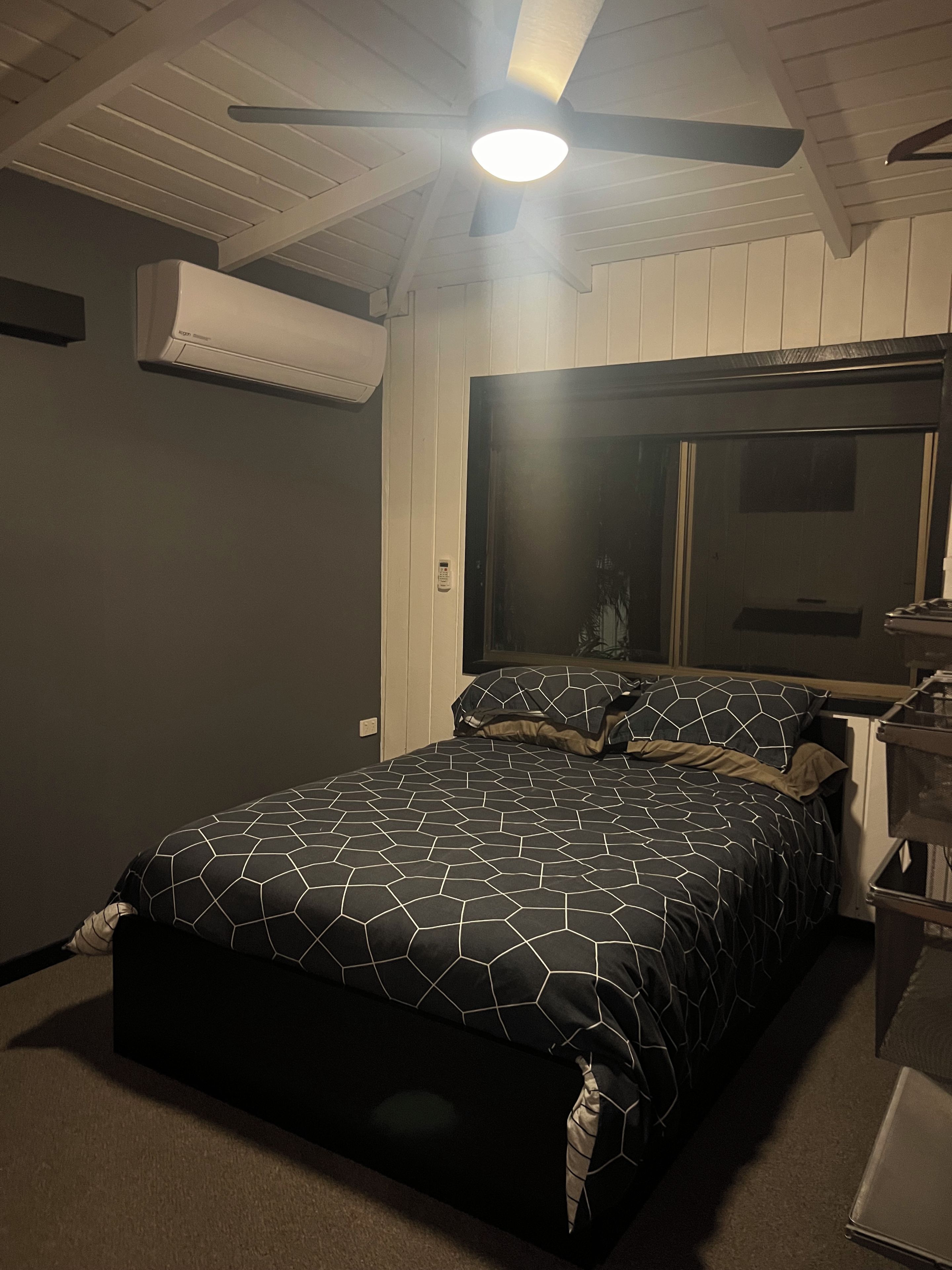 A bedroom with a bed, a ceiling fan, an air conditioning unit, a window, and a nightstand.