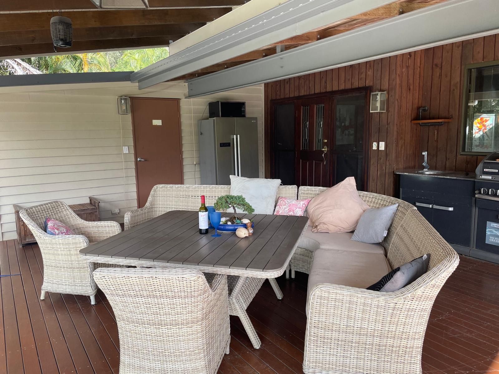 Un patio exterior con mobiliario de mimbre, una mesa con una botella y una planta, y una zona de cocina con una parrilla.