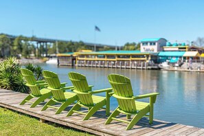 A wooden dock with green chairs facing a calm body of water, a bridge in the background, and a building with a blue roof. - Summersalt Retreat• Canal Views • Walk to Dining • Pet Friendly (Gulf Shores)