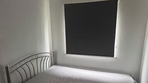 Un lit simple avec un cadre en métal et un matelas ordinaire, dans une pièce dotée d’une fenêtre.