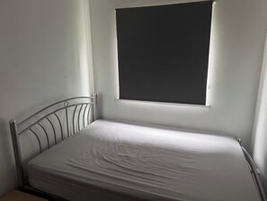 A single bed with a metal frame and a plain mattress in a room with a window. - Apartment Sleeps 4 with Parking (London)