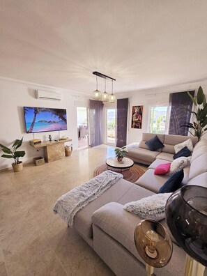 Een moderne woonkamer met een grote flatscreen-tv, een zithoek, een salontafel en een eethoek zichtbaar door een glazen deur.
