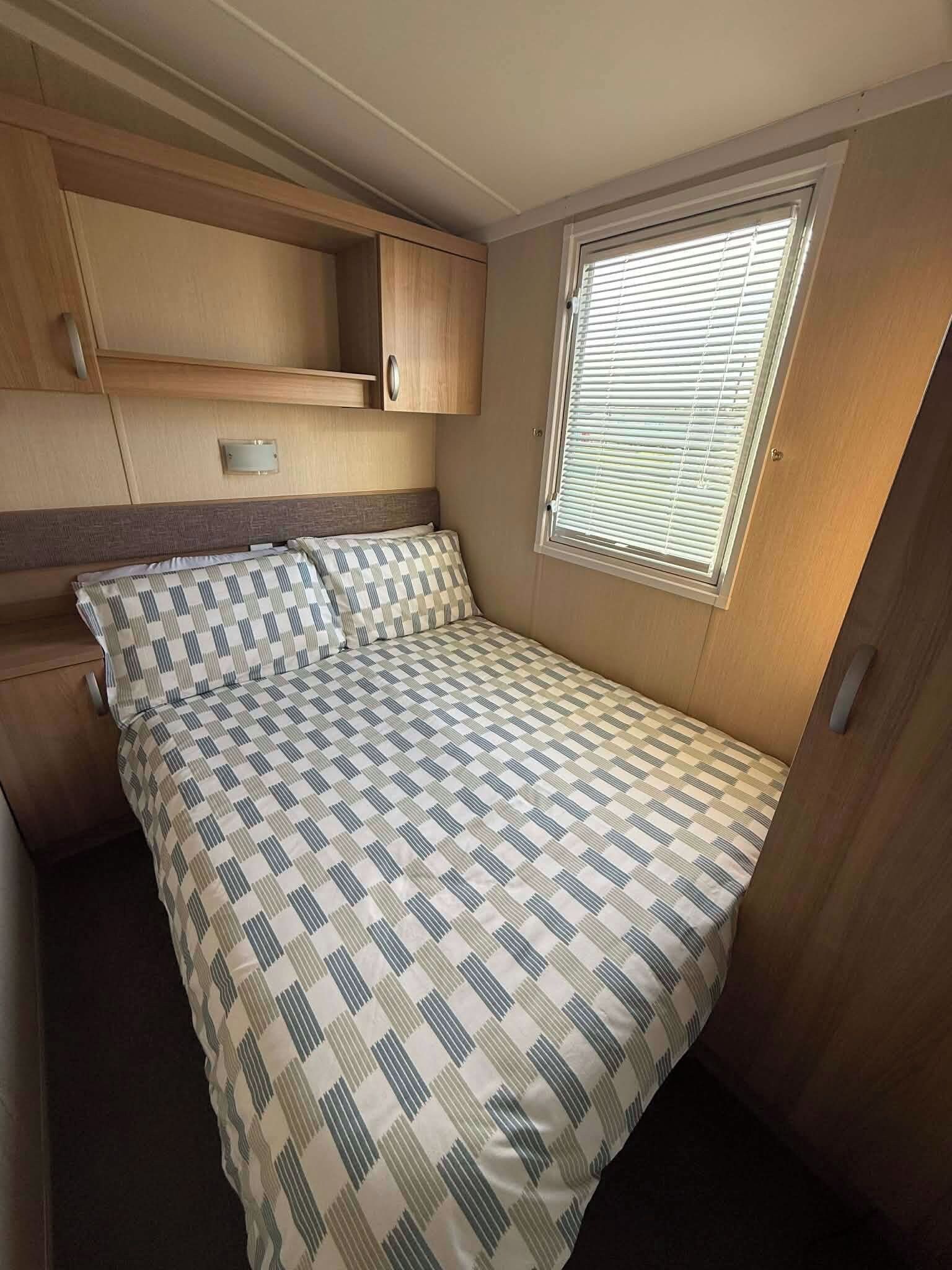 Une chambre compacte dans un véhicule récréatif, comprenant un lit, une fenêtre avec stores et des armoires intégrées en bois.