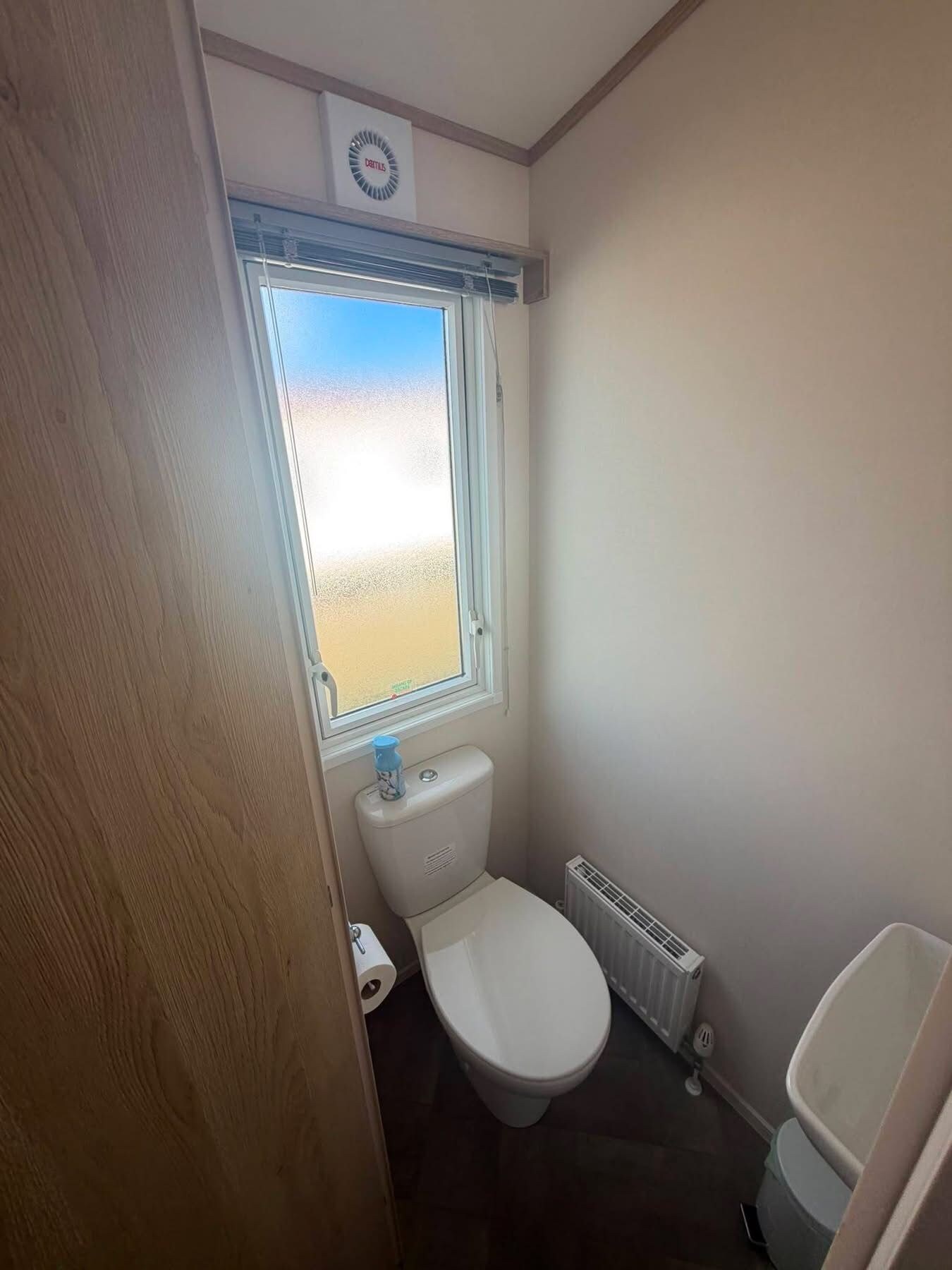 A small bathroom with a toilet, a radiator, and a window with a view of the beach.