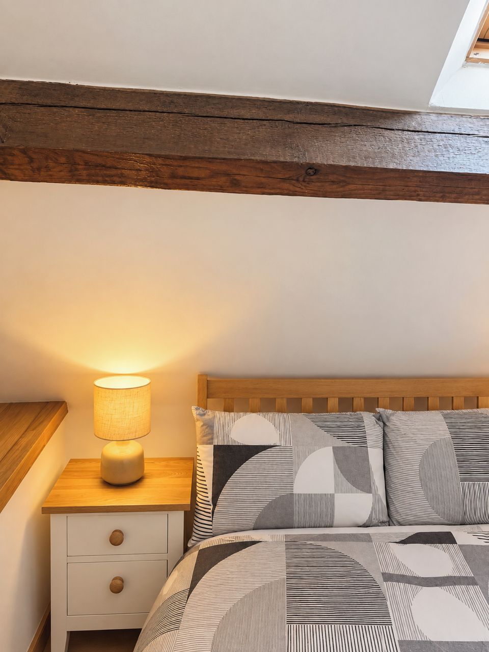 Een slaapkamer met een houten hoofdbord, een bedlampje, een bed met beddengoed met een patroon, en een witte nachtkastje met lades.