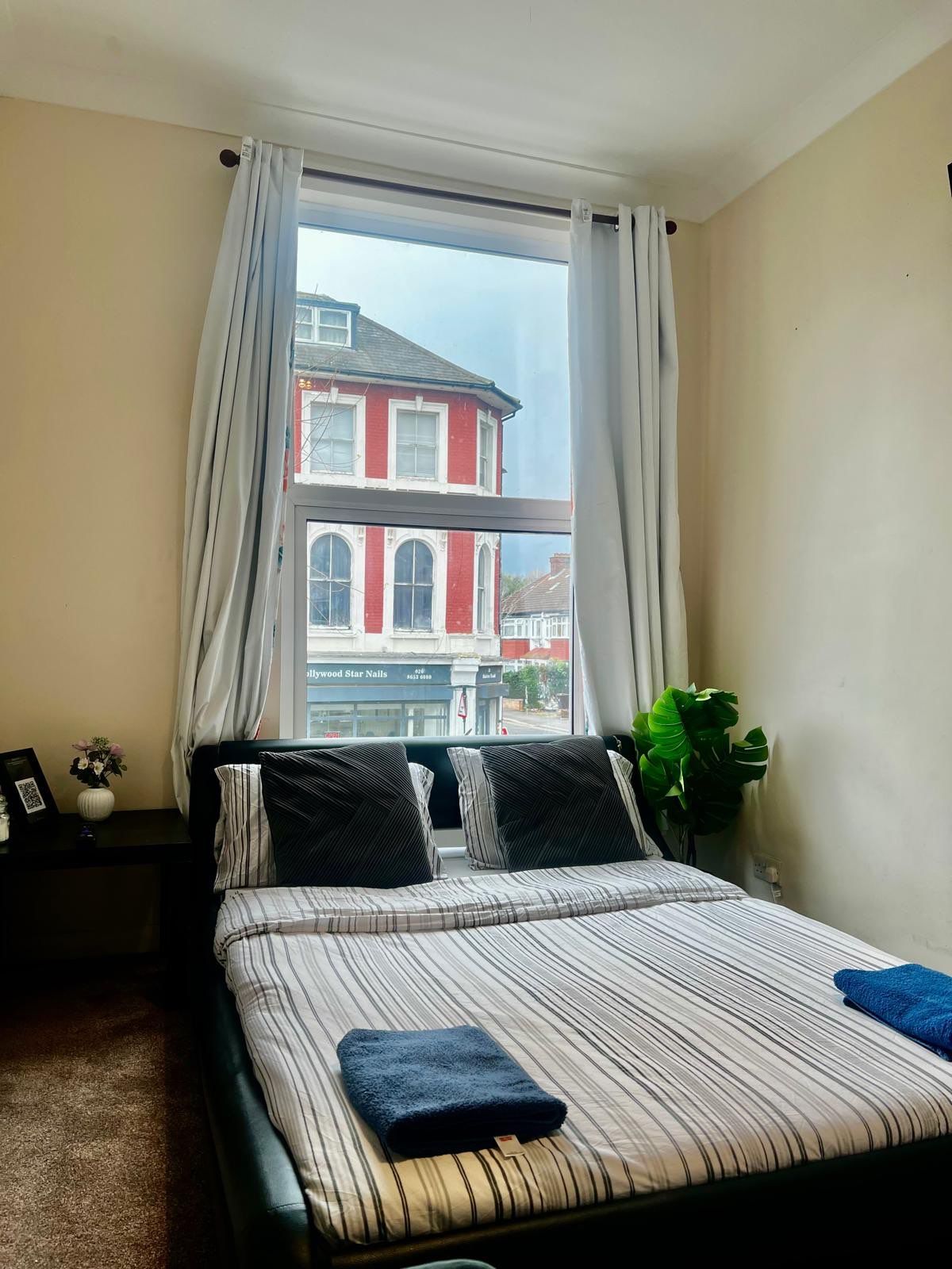 A bedroom with a large window, a bed with striped bedding, a nightstand with a vase and picture frame, and a view of a red building.
