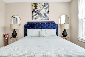 A bedroom with a tufted headboard, two bedside tables with lamps, a large abstract painting above the bed, and a window with blinds.