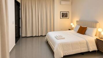 Habitación de hotel con una cama, una mesita de noche con lámpara, una puerta y un ventanal con cortinas.