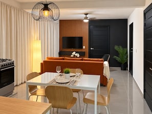 A modern living room with a sofa, dining table, and chairs. There is a flat-screen TV mounted on the wall, a ceiling fan, and a potted plant.