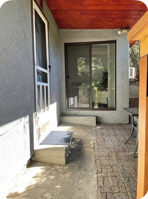 Ein überdachter Patio-Bereich mit Betonboden, Holzboden und einer Glastür, die in einen Garten führt.