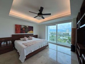 A bedroom with a bed, a ceiling fan, a large window with a city view, and artwork on the wall. - Shangri La - Condominio de Lujo Frente al Mar (Puerto Vallarta)