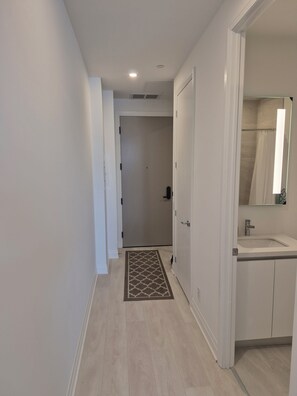 A hallway with a door leading to a bathroom, a patterned rug, and a light-colored floor. - Brand New Studio on LRT Line (Toronto)