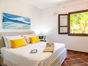 Une chambre à coucher avec un lit, des oreillers, un tableau, une table de chevet, une lampe et une fenêtre donnant sur des arbres.
