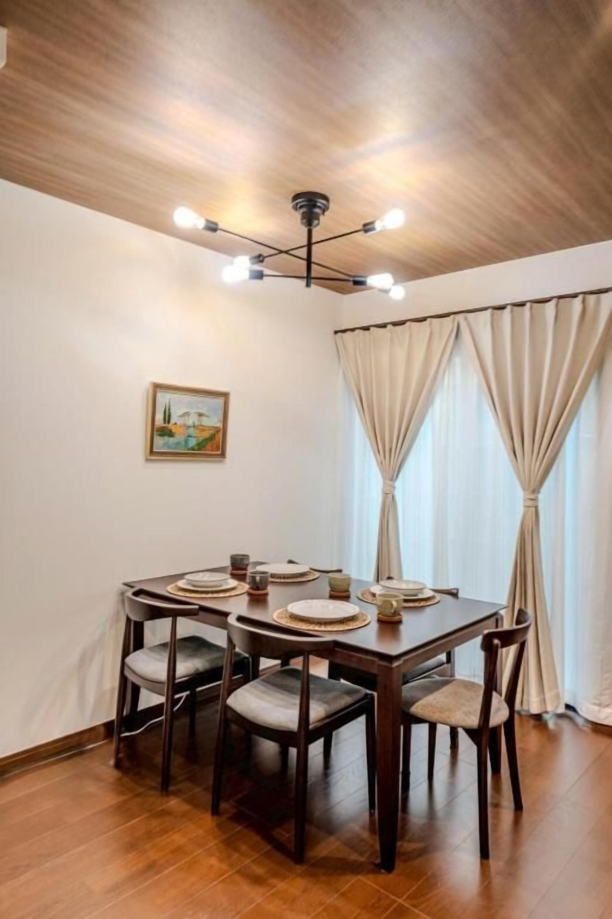 A dining area with a wooden table set for four, a ceiling light fixture, and a window with curtains.
