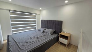 Un dormitorio con una cama, una mesita de noche, una ventana con persianas y un enchufe.