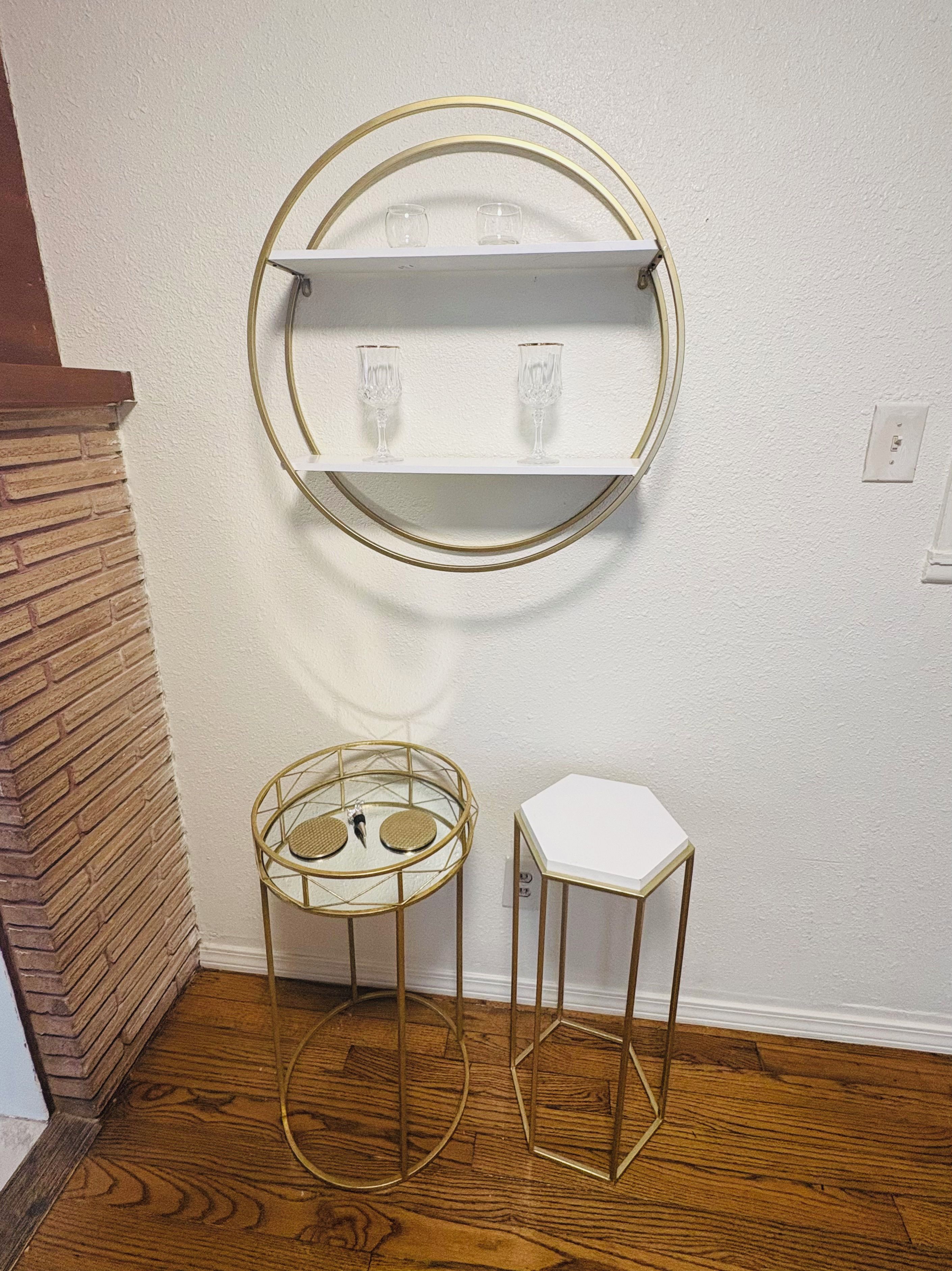 Une pièce avec un mur blanc, une étagère ronde à cadre doré sur laquelle reposent deux verres, une petite table ronde à cadre doré et un tabouret blanc.