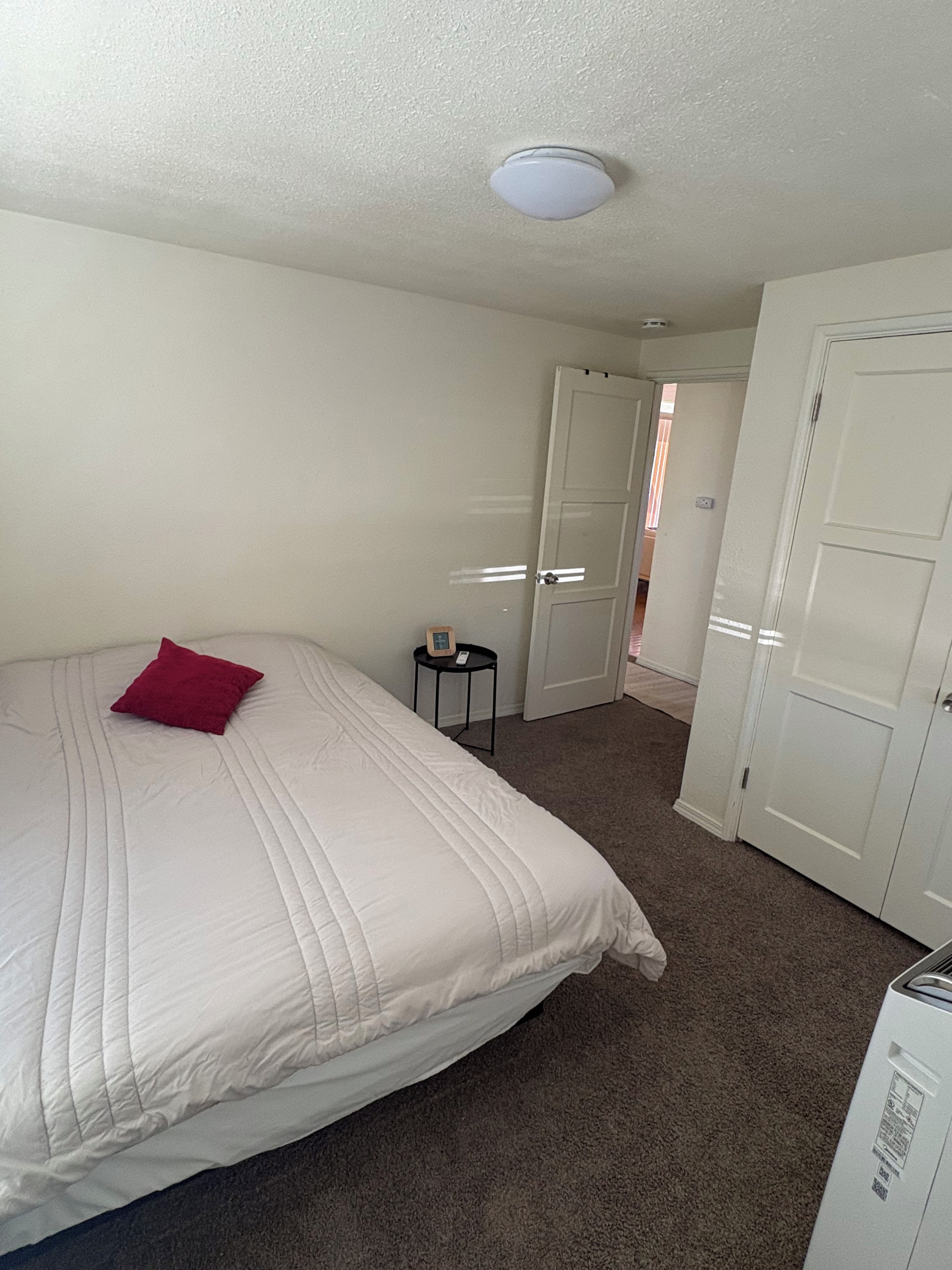 A bedroom with a bed, a small bedside table, a red pillow, and a white door.
