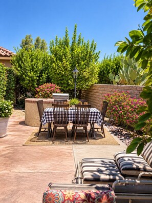 Um pátio externo com mesa e cadeiras, churrasqueira e diversas plantas e flores.