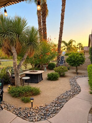 A patio area with a fire pit, seating, and a variety of plants including palm trees and shrubs. - Redford Villa- 3BR Home + 1BR Private Casita, Pool/Spa, Close to Polo (Indio)