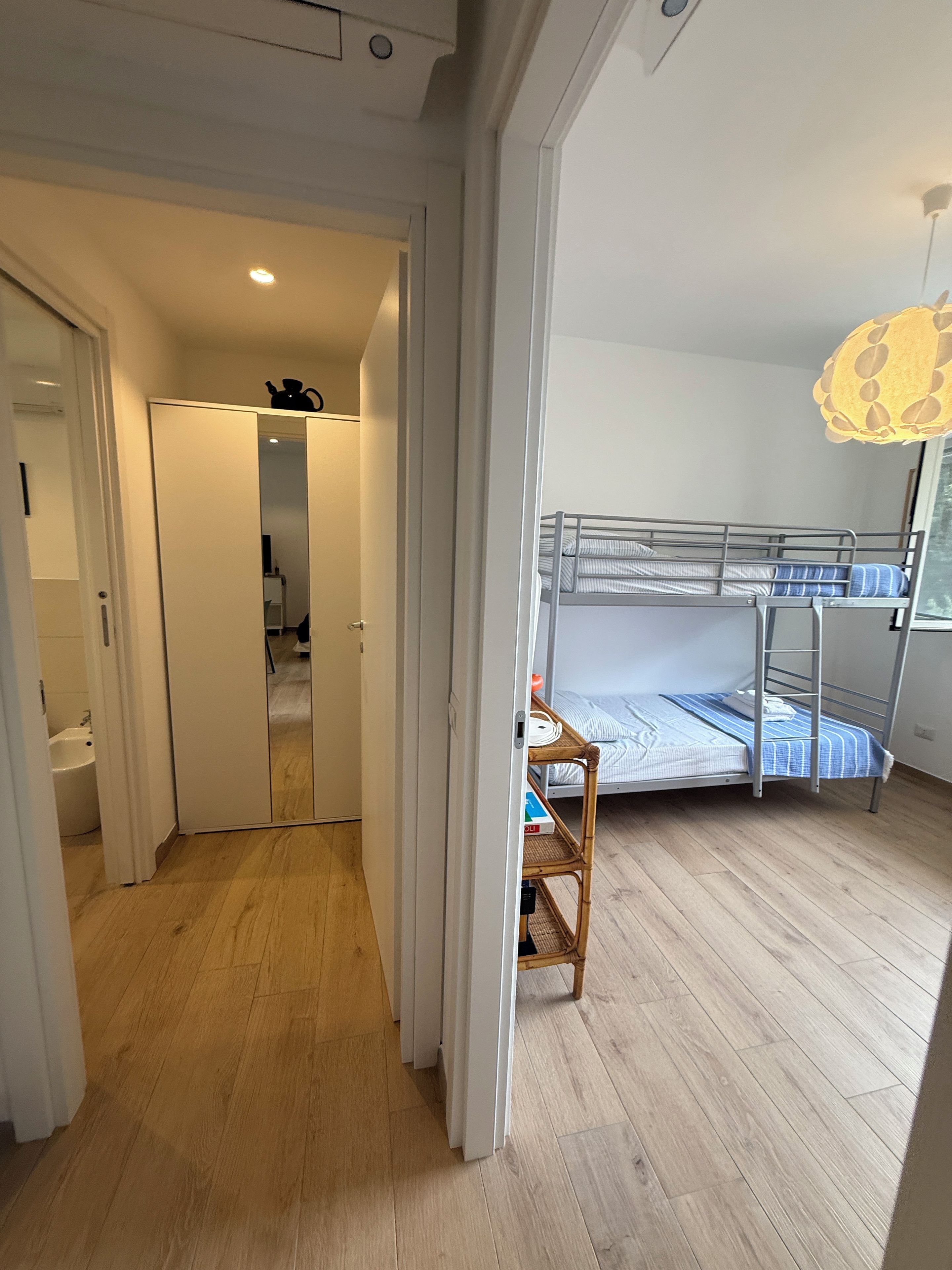 Een kamer met een stapelbed, een houten vloer, een witte garderobe en een badkamer met een wit wastafelmeubel.