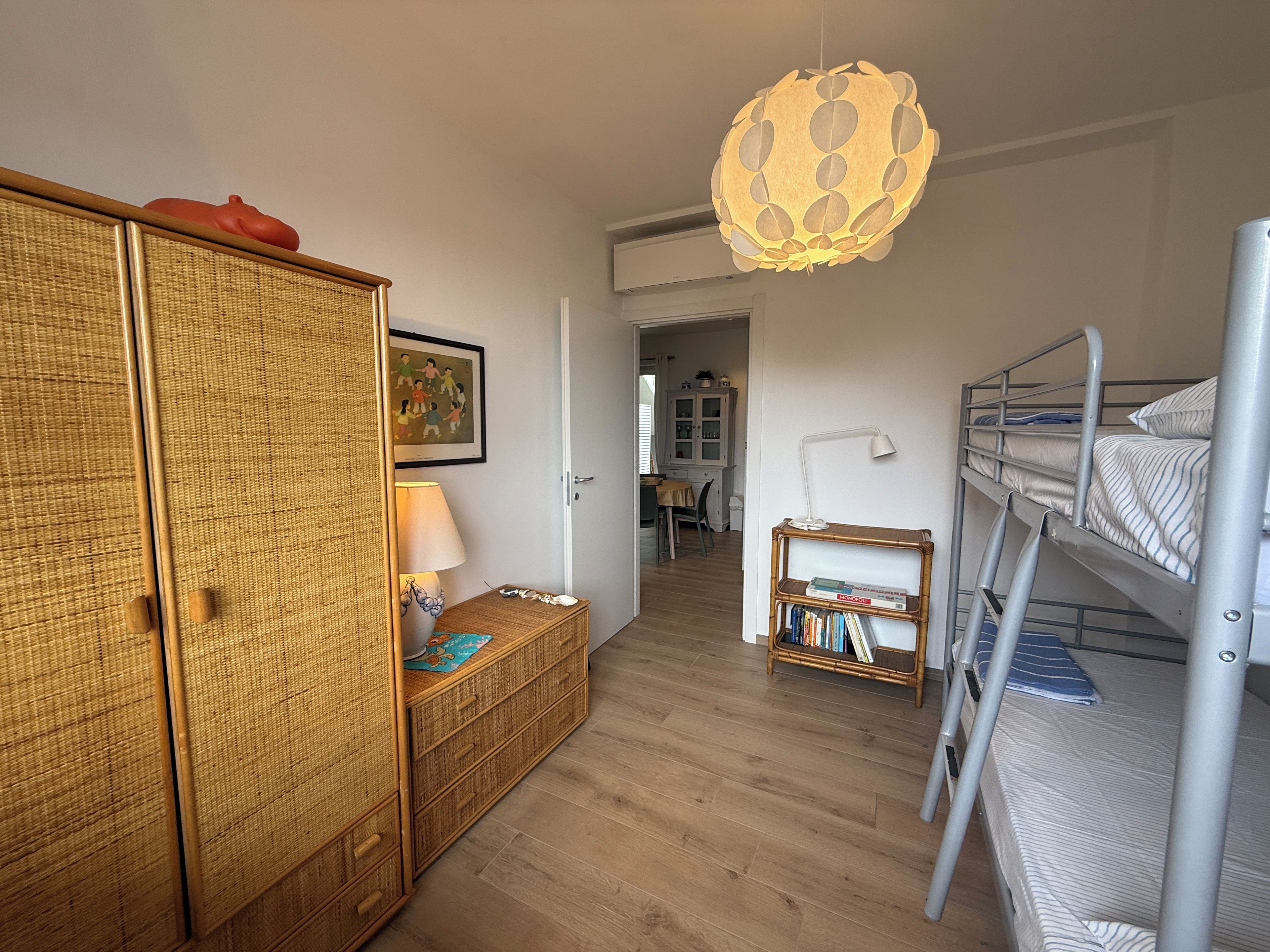 A room with a bunk bed, a wicker wardrobe, a lamp, and a wall-mounted shelf with books.
