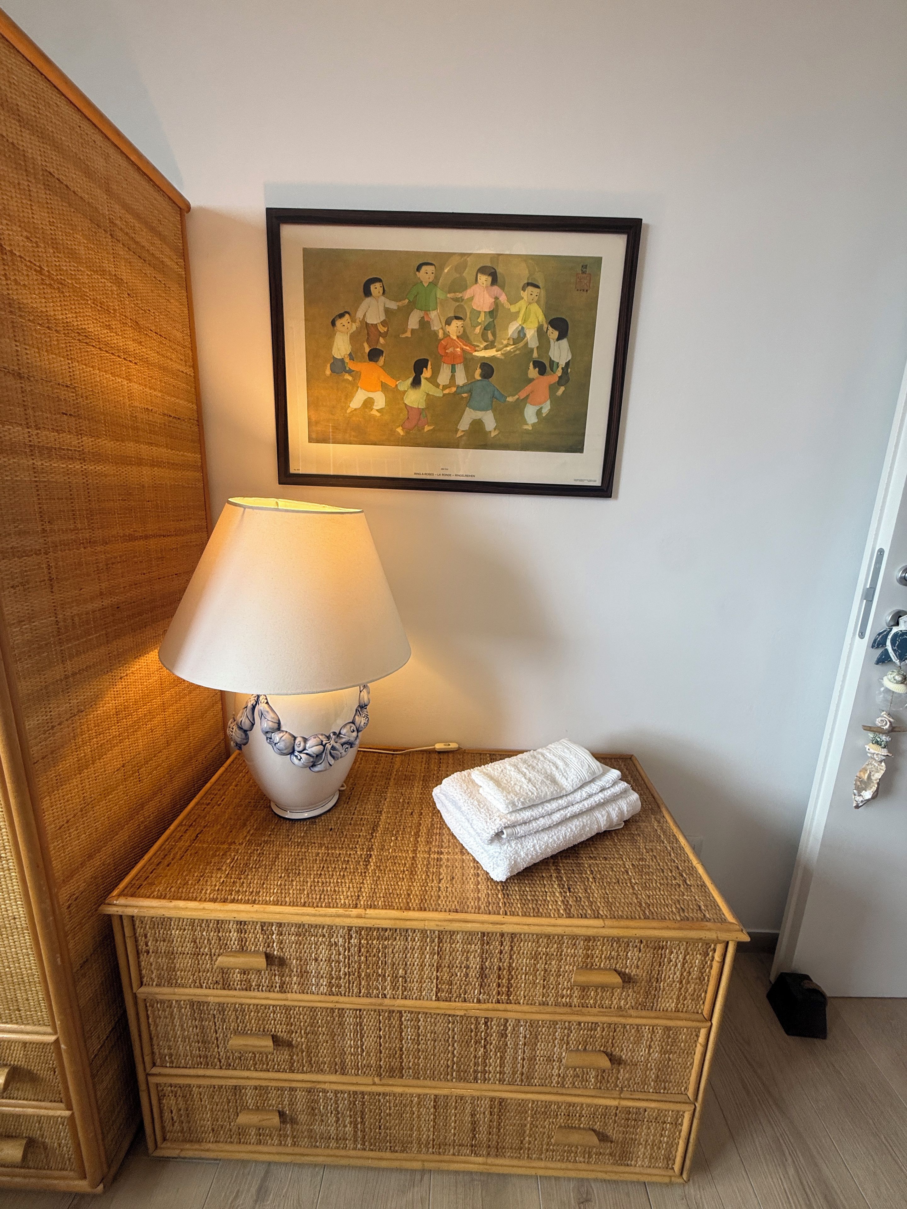 Quarto com abajur, uma obra de arte emoldurada, cômoda de palha e toalha dobrada com cuidado.