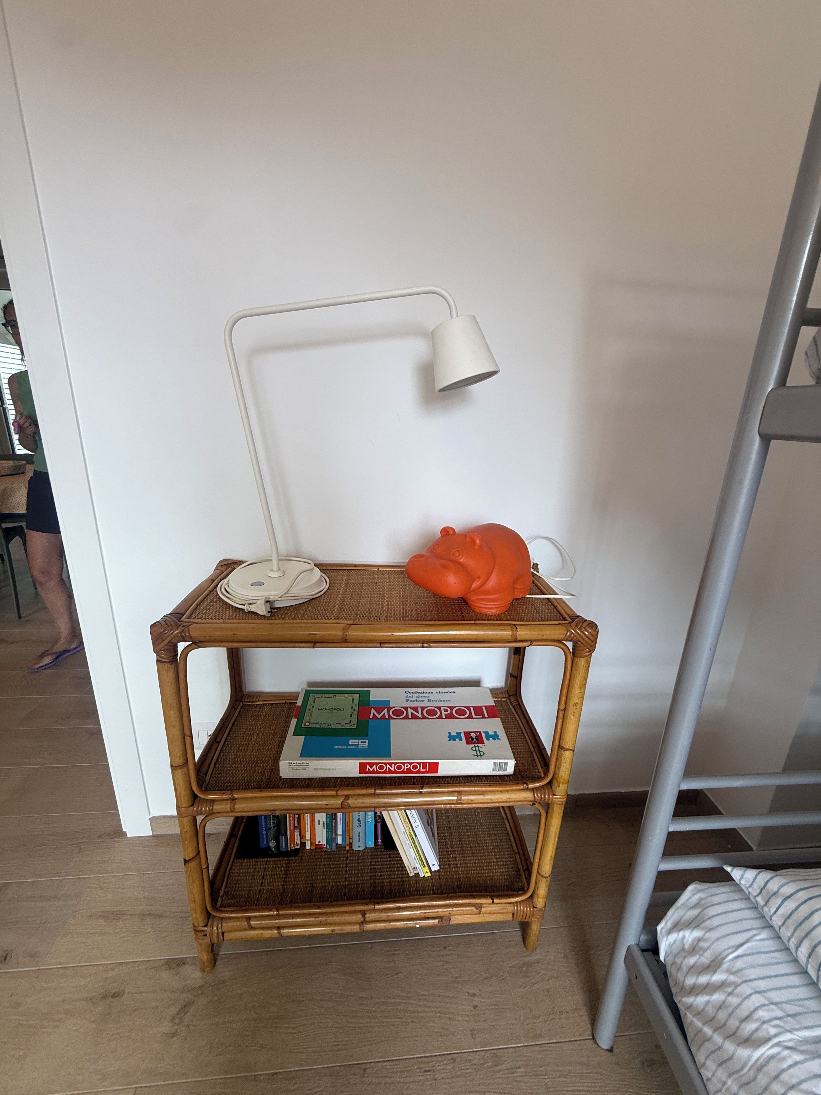 A room with a white wall, a white desk lamp, a wicker side table with a Monopoly board game, and a metal bunk bed.