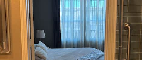 A hotel room with a bed, a bedside lamp, and a view of the outside through sheer curtains.