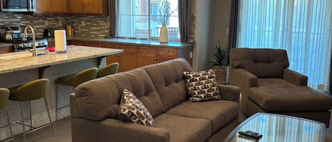 A modern living room with a sofa, armchairs, a coffee table, and a kitchen area with a microwave.
