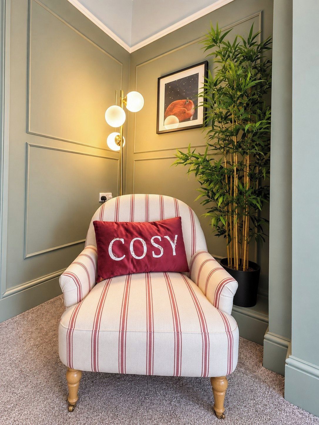 Una habitación de esquina con un sillón a rayas, un cojín rojo con la palabra 'COSY', una lámpara de pared, un cuadro enmarco y una planta de bambú.