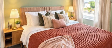 A bedroom with a large bed, two bedside tables, a hanging light fixture, a window with curtains, and a patterned rug.