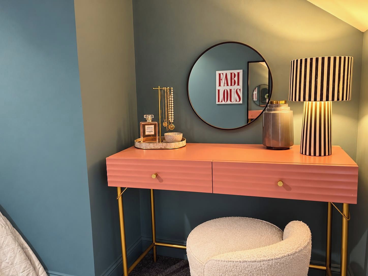 A pink vanity with a gold frame, a round mirror, a lamp, and a small stool.