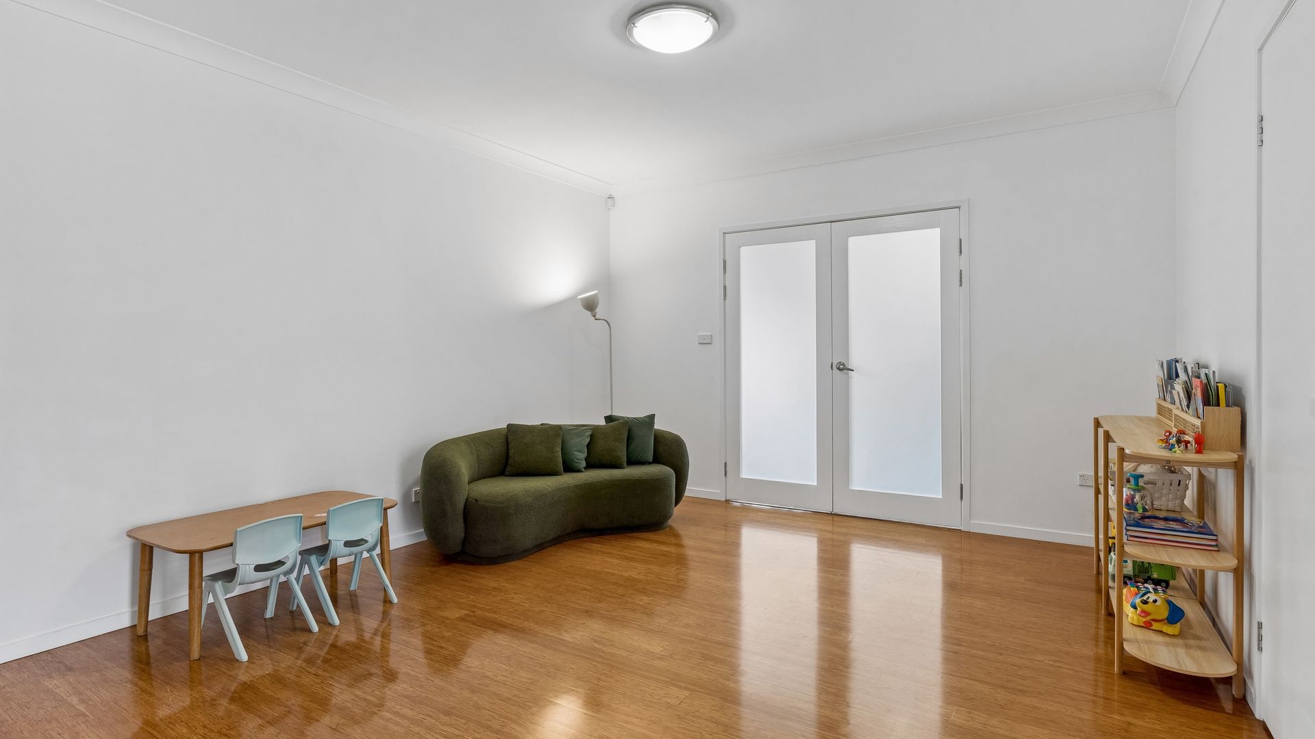 A spacious room with a wooden floor, a green sofa, a small table with chairs, and a bookshelf with children's books.