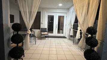 A covered outdoor area with a tiled floor, white curtains, and potted topiary plants.