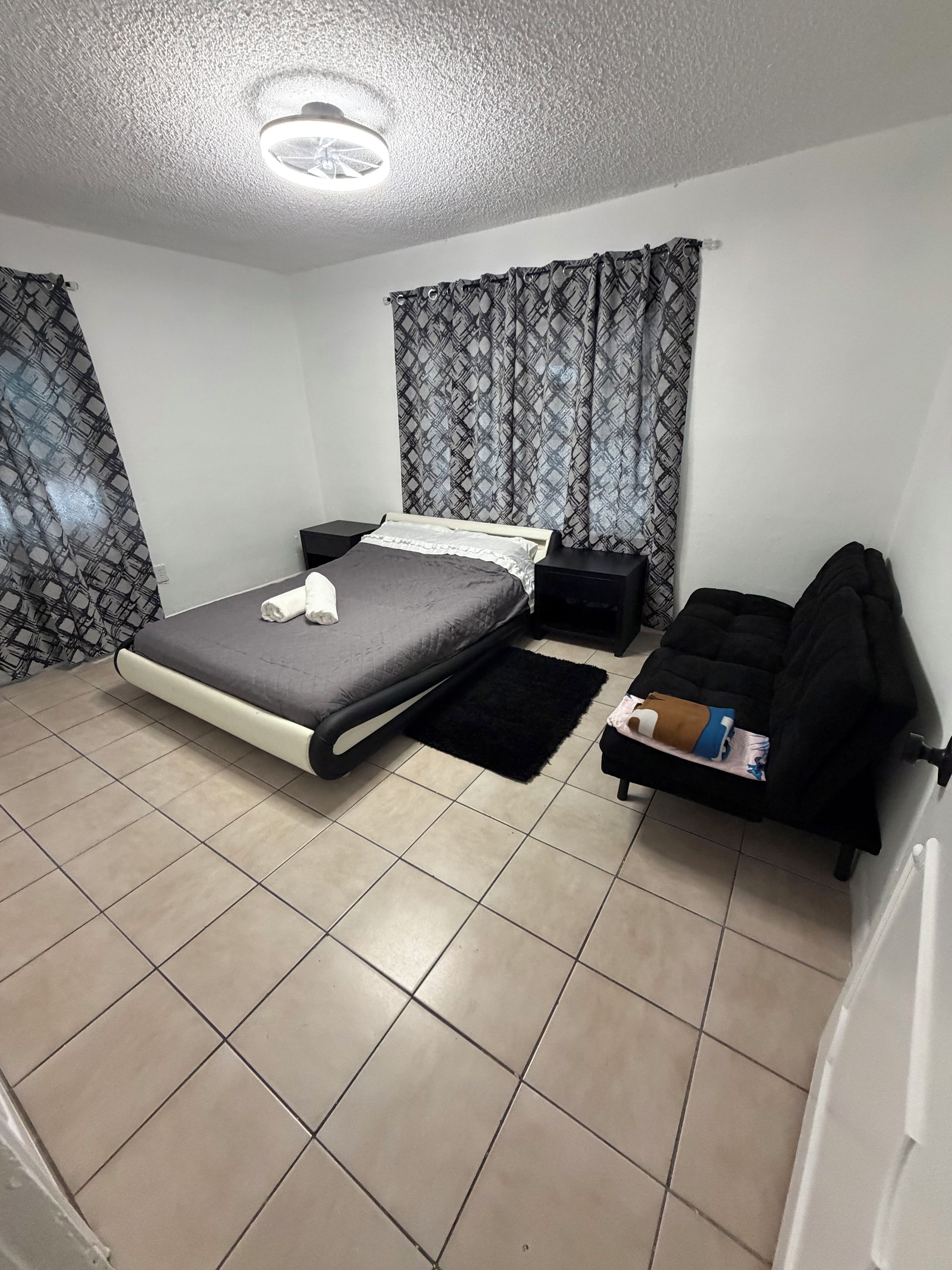 A bedroom with a bed, a bedside table, a black sofa, a black rug, and patterned curtains.