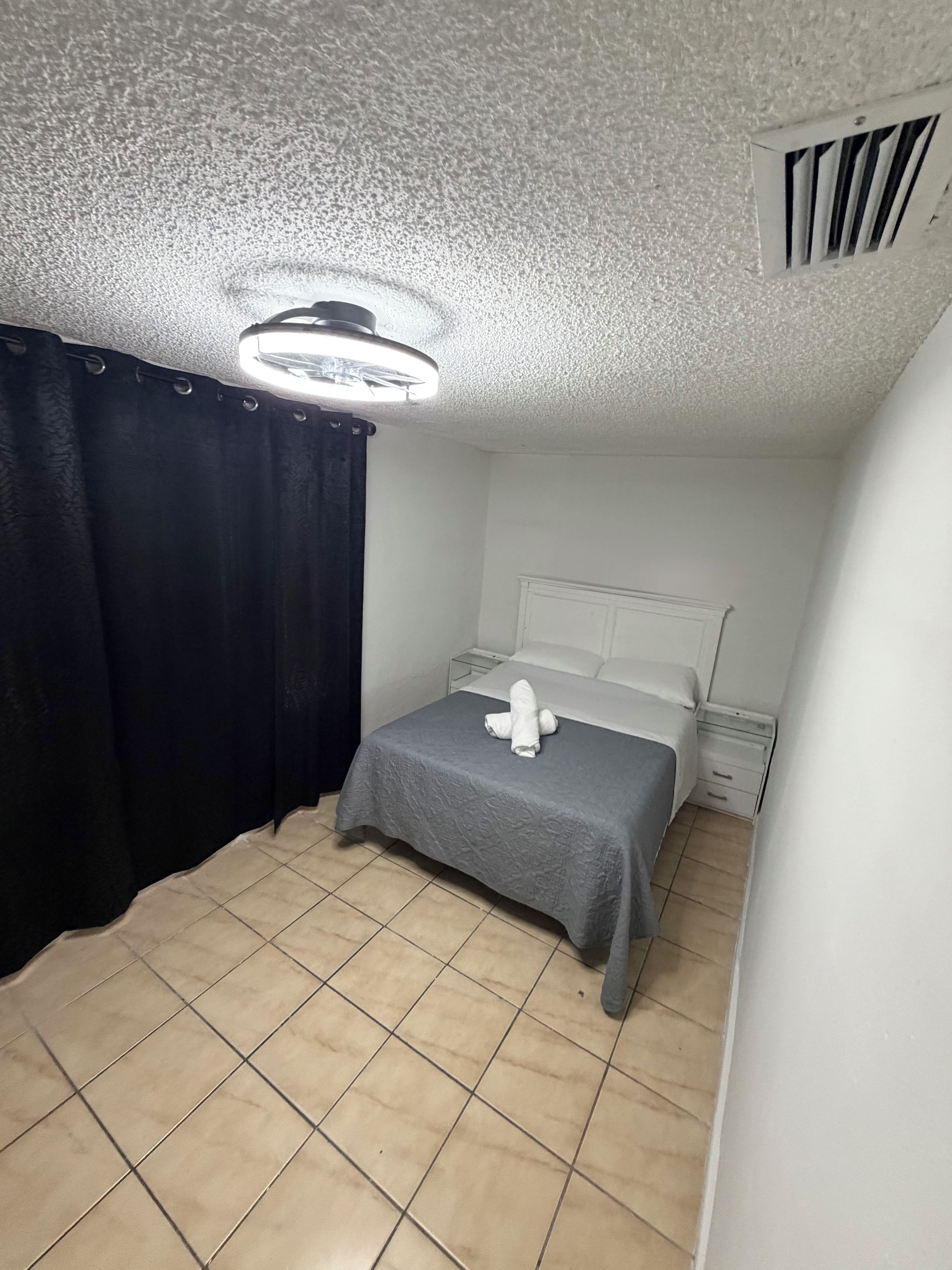 Une petite chambre avec un lit simple, recouverte d’une courtepointe grise, un oreiller blanc et une serviette blanche posée sur le lit. On y trouve une table de chevet blanche avec une lampe, un rideau noir et une bouche d’aération au plafond.