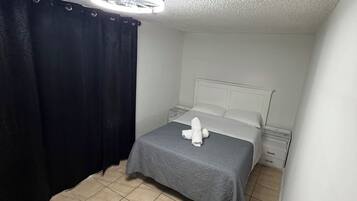 Une petite chambre avec un lit simple, recouverte d’une courtepointe grise, un oreiller blanc et une serviette blanche posée sur le lit. On y trouve une table de chevet blanche avec une lampe, un rideau noir et une bouche d’aération au plafond.