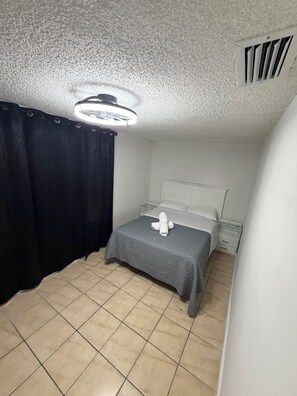 A small, single-bed room with a grey bedspread, a white pillow, and a white towel on the bed. There is a white nightstand with a lamp, a black curtain, and a ceiling vent.