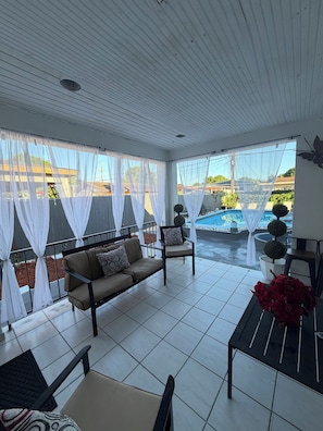 A covered patio area with a sofa, chairs, a table, and a view of a swimming pool.