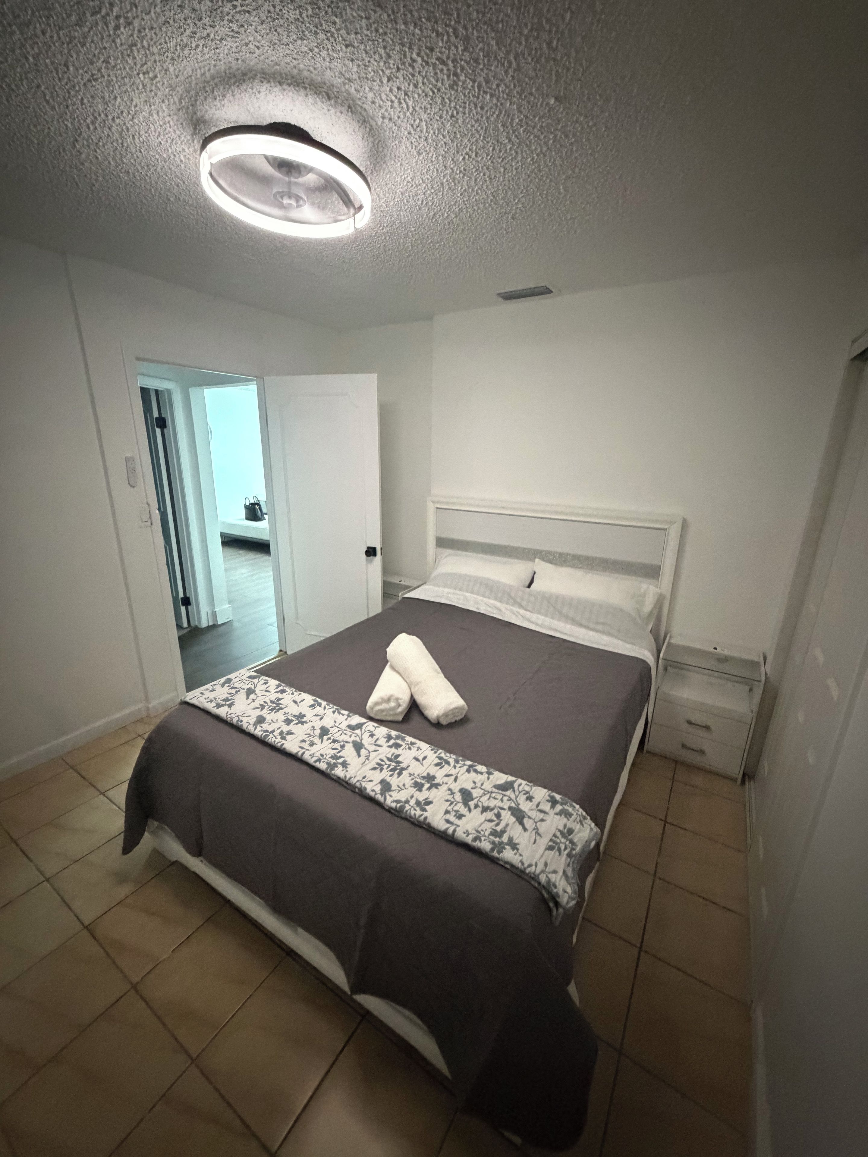 Un dormitorio con cama, una mesita de noche, una puerta a otra habitación y una lámpara en el techo.