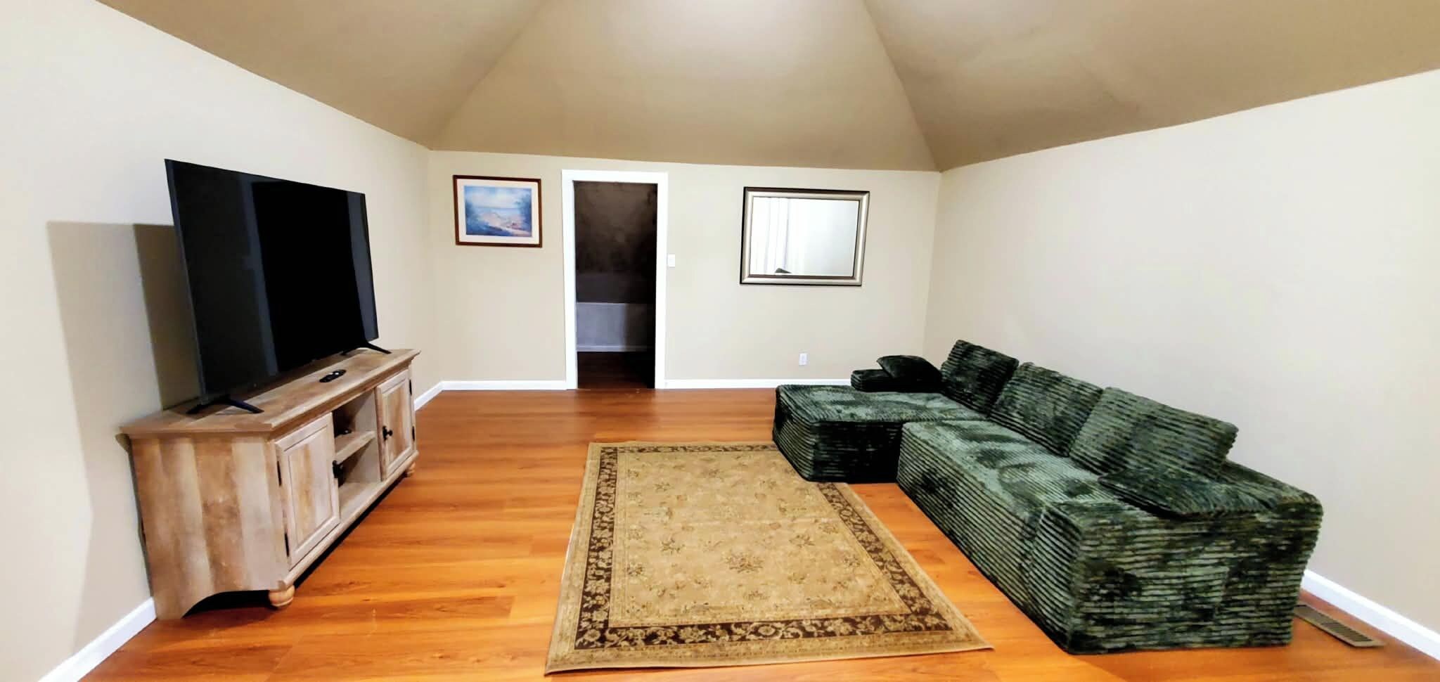 A living room with a flat-screen TV on a wooden stand, a green sofa, a patterned rug, and two framed pictures on the wall.