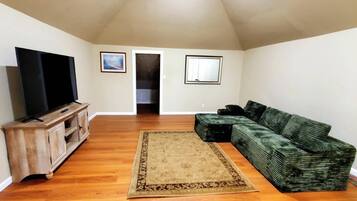 A living room with a flat-screen TV on a wooden stand, a green sofa, a patterned rug, and two framed pictures on the wall.