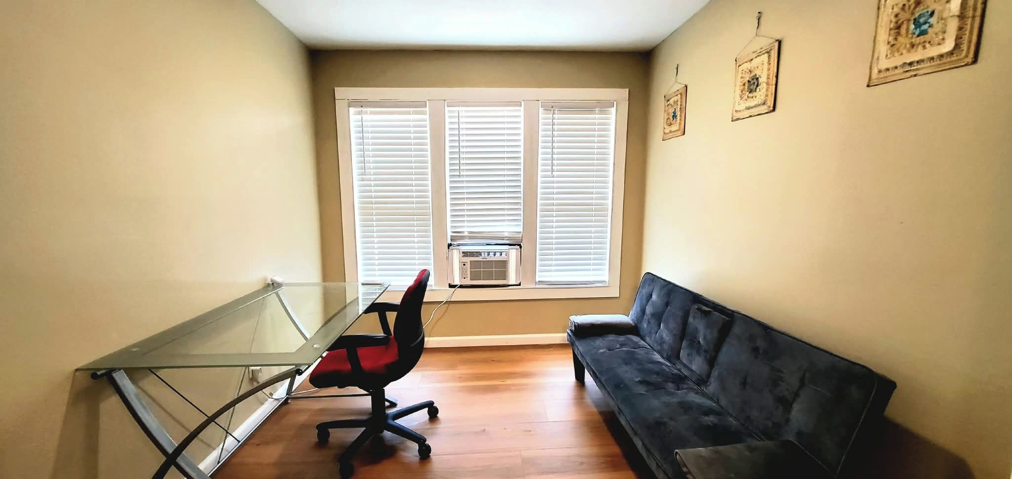 Une pièce avec un bureau à plateau en verre, un fauteuil de bureau rouge, un canapé noir et une fenêtre avec des stores.