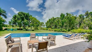 Zona de piscina con sillones de descanso y una mesa, rodeada de exuberante vegetación y árboles.