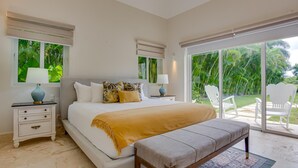 Un dormitorio con una cama grande, dos mesitas de noche, un banco y vistas a un jardín tropical a través del ventanal.