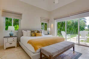 Een slaapkamer met een groot bed, twee nachtkastjes, een bankje en uitzicht op een tropische tuin door de glazen deur.